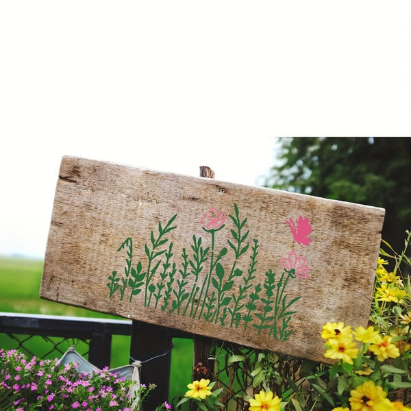 A rustic rectangular wooden sign painted with green plants and pink flowers, shown outdoors in a garden setting with yellow flowers and greenery in the background.