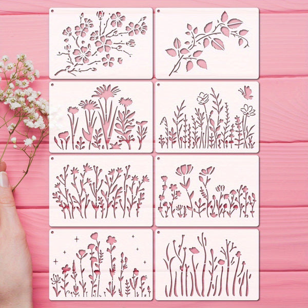 Six square metal stencils with pink floral and botanical cutout designs arranged in a 3x2 grid on a pink wooden surface, with a hand holding baby's breath flowers at the left edge.