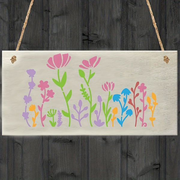 A rectangular white wooden hanging plaque with a colorful floral design, suspended by twine against a dark gray wood plank background.