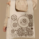A beige canvas tote bag with a brown steampunk gear pattern, held by a person wearing a matching beige hoodie against a solid beige background.