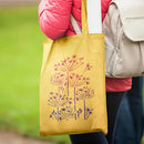 A person wearing a pink puffer jacket carries a mustard yellow reusable tote bag with a stylized dandelion print in dark purple. The person is also wearing a light beige backpack, set against a blurred green outdoor background.