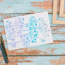A white art print featuring stylized botanical dandelion-like patterns in blue, purple, and gray, displayed on a rustic distressed wooden table alongside paintbrushes and picture frames.