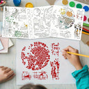 A top-down view of a hand coloring a stencil design on a white sheet of paper with a yellow pencil, with three unfolded white stencils showing various city and abstract patterns. The scene is set on a light-colored wooden desk surrounded by paint palettes, water bottles, and paint tubes.