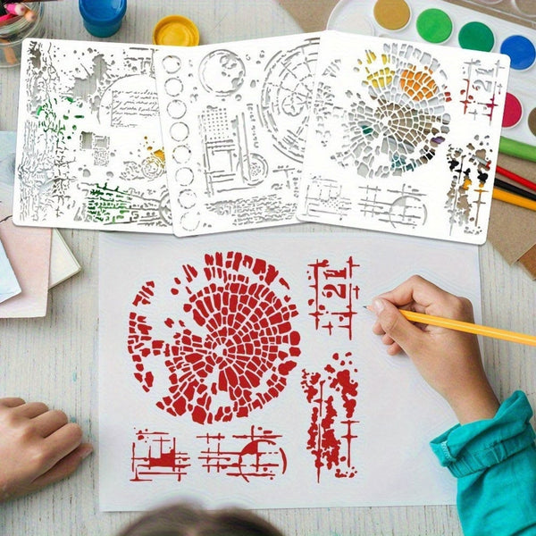A top-down view of a hand coloring a stencil design on a white sheet of paper with a yellow pencil, with three unfolded white stencils showing various city and abstract patterns. The scene is set on a light-colored wooden desk surrounded by paint palettes, water bottles, and paint tubes.