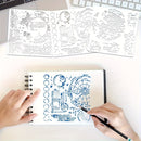 A top-down view of a person's hands using a blue ink stamper on a spiral notebook, with two white stencil sheets featuring celestial and geometric designs laid out on a white desk above.