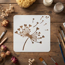 A square white stencil with a dandelion design laid flat on a rustic wooden table, surrounded by paintbrushes, dried flowers, a sponge, and small jars of cream-colored substance.