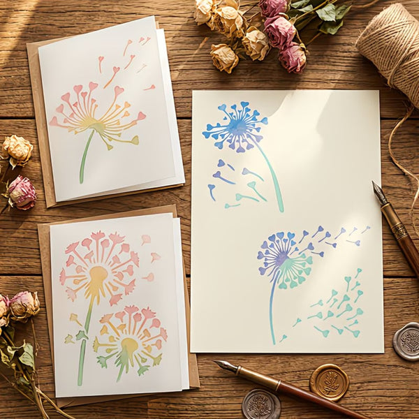 Three greeting cards with dandelion illustrations are arranged on a rustic wooden table with dried roses, twine, pens, and wax seals. The cards feature pastel watercolor-style dandelion designs in pink, yellow, blue, and green on white cardstock.