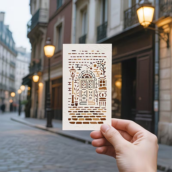 A hand holds a small rectangular card featuring a laser-cut architectural street scene with a central arched doorway. The background shows a softly blurred cobblestone street and old buildings with lit street lamps.