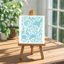 A square art print with a white floral lace pattern on a light blue background, displayed on a small wooden easel on a wooden table in a bright room with a window and green plants in the background.