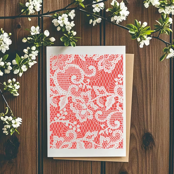 A square greeting card with a coral pink and white lace pattern, displayed on top of a kraft envelope on a dark wood background with white blossoms.
