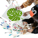 Two children painting tropical monstera leaves on white canvases with markers, surrounded by a scattered collection of colorful markers on a white background.