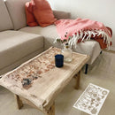 Beige sectional sofa with pink cushions and blanket, wooden coffee table with decorative items, and floral patterned mat on floor.