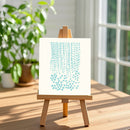 A small wooden easel stands on a sunlit wooden table, holding a white art print or canvas with a light blue abstract pattern. The background shows a softly blurred indoor setting with a window and potted plants.