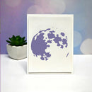 A minimalist art print featuring a purple silhouette of the moon on an off-white card, displayed on a white surface with a small potted succulent and a purple and blue bokeh background.