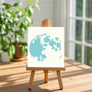 A small wooden easel stands on a wooden table displaying a square canvas with a light blue abstract globe or map-like graphic on a white background. The scene is softly lit by a window, with blurred green plants in the background.
