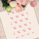 A square cream-colored card or paper featuring a repeating pattern of small pink fish and dots, styled flat-lay with pink roses and green leaves at the top edge on a light fabric background.