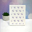 A white rectangular card or print featuring a repeating pattern of small purple fish and dots, standing upright on a white surface. A small potted succulent sits to the left, and the background is a soft pastel purple and blue bokeh-style backdrop.