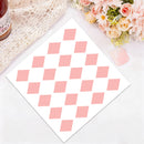 A square white card or sheet featuring a repeating pink diamond pattern, photographed on a light surface with decorative props including lace, flowers, and a bottle edge.