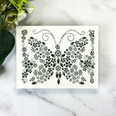 A rectangular white card or stencil with a black butterfly design made of floral and leaf motifs, placed on a white marble surface with green leaves and white flowers as props.