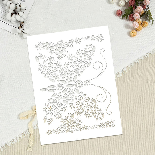 A white rectangular card or stencil with an intricate laser-cut floral and scroll pattern, photographed on a light fabric surface with cotton stems and small flowers as styled props.