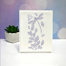 A vertical white art card featuring a lavender line drawing of a dragonfly, leaves, and small flowers, standing on a white surface with a small potted succulent and a purple-toned bokeh background.