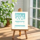 A small wooden easel stands on a wooden table in a bright room, holding a white square canvas with a light blue abstract dripping pattern. A blurred green plant and a window are in the background.