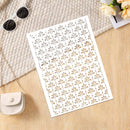 A white rectangular panel with an allover laser-cut leaf or fan pattern, photographed on a light wood surface with a beige knit fabric, sunglasses, a small cream purse, and flowers as styling props.