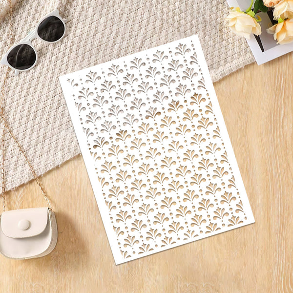 A white rectangular panel with an allover laser-cut leaf or fan pattern, photographed on a light wood surface with a beige knit fabric, sunglasses, a small cream purse, and flowers as styling props.