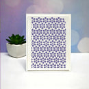 A white square art print or card featuring a repeating purple floral pattern, standing upright on a white surface with a small potted succulent and a soft pastel bokeh background.