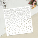 A square white laser-cut stencil or craft template with irregular circular cutouts, displayed on a textured beige fabric background with decorative cotton and dried flowers.