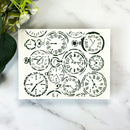 A square white art print or card featuring a collage of black vintage pocket watch illustrations, displayed on a white marble surface with green leaves and white flowers in the corner.