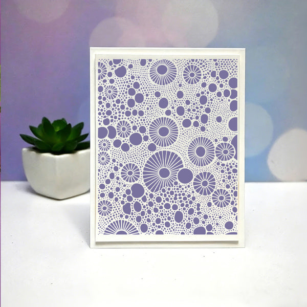 A square white card with a purple geometric dot and circle pattern, standing on a white surface with a small potted succulent in the background against a soft purple and blue bokeh backdrop.