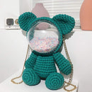 A crocheted teddy bear-shaped shoulder bag in teal green, featuring a clear circular clear plastic dome filled with pastel gumballs and a gold chain strap, displayed on a white shelf.