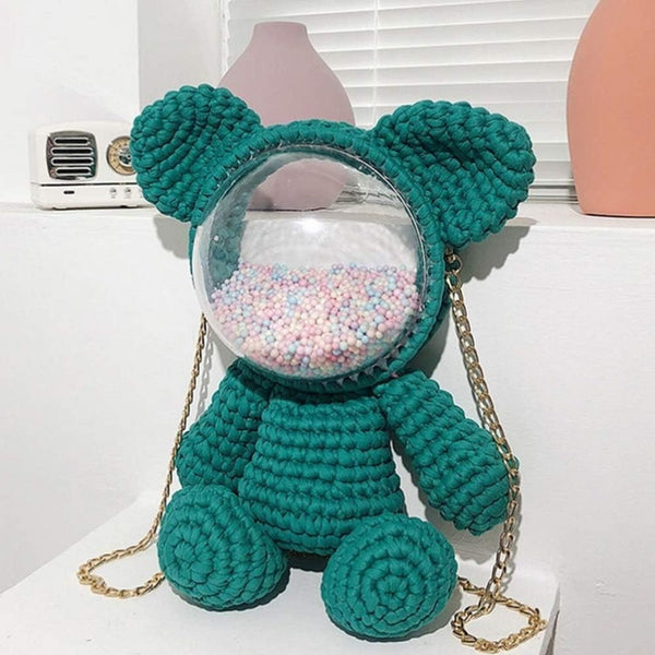 A crocheted teddy bear-shaped shoulder bag in teal green, featuring a clear circular clear plastic dome filled with pastel gumballs and a gold chain strap, displayed on a white shelf.
