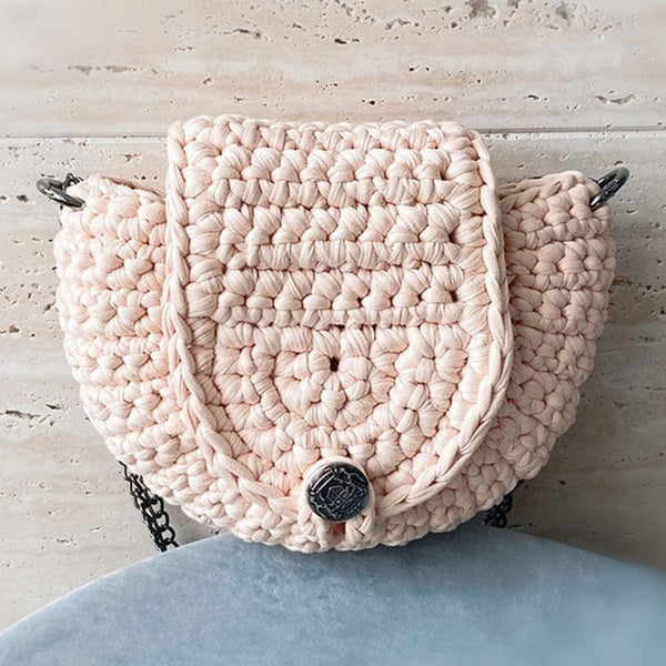 A handmade, chunky crocheted shoulder bag in a light blush pink color, featuring a central button closure and a textured woven body, resting on a light gray surface.