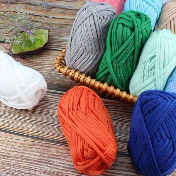 Multiple thick yarn balls in assorted colors arranged on a rustic wooden surface, including white, gray, green, mint, peach, orange, and blue. Some yarn balls are placed in a small woven basket, and a decorative green stone is visible in the background.