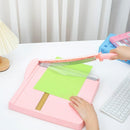 Person using a pink paper trimmer on a white desk with a keyboard and lamp in the background.
