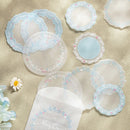 A set of pastel blue and white floral lace-edged round and square coasters arranged on a beige textured surface, with a matching translucent envelope at the bottom and a small daisy flower prop on the left.