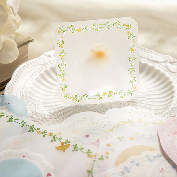 A square translucent white card or coaster with a delicate green vine and yellow flower border, standing upright on a white scalloped plate. Other matching decorative pieces are scattered around on a soft beige surface.