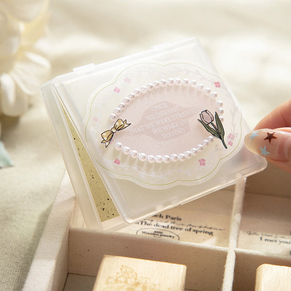 A hand with decorated nails holds a clear plastic stamp box containing a decorative oval stamp with a pearl border, tulip illustration, and the phrase 'We dream that all we aspire to, we wake up to again.' The box is shown over a beige stamp set in soft, warm lighting.