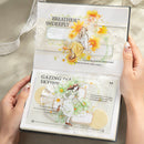 A person holds open a decorative book or journal showing two illustrated spread pages with floral and whimsical illustrations, lace overlays, and printed text, photographed against a soft neutral fabric background.