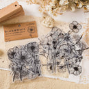 Black and white floral sticker set with clear vinyl sheets showing detailed line-art flowers and leaves, displayed on a textured cream surface with a wooden stamp block and dried flowers as props.