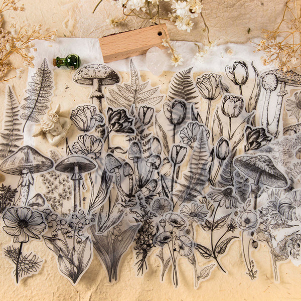 A sheet of clear stamps or decals featuring black and gray botanical line art, including mushrooms, ferns, tulips, and wildflowers, arranged on a textured beige surface with dried flowers and a small wooden tag.