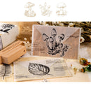 A styled flat-lay of a clear acrylic stamp with a black mushroom illustration, shown with a wooden block, paper, and decorative props on a light beige surface. A white banner at the top shows three additional mushroom stamp designs.