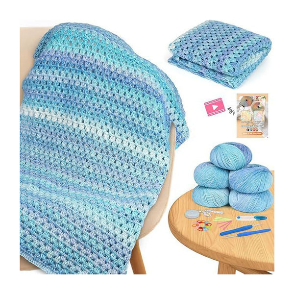 A crocheted blanket in variegated light blue, aqua, and white yarn draped over a chair, with folded matching yarn shown at top right and a round wooden table at bottom right holding several blue yarn balls, crochet hooks, needles, and tools. A small video icon and booklet are also visible.