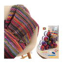 A multicolored crocheted blanket draped over a beige chair, shown alongside a round table holding yarn balls, a crochet kit, and a booklet titled 'Learn to Crochet Crocheting Striped Blankets'.