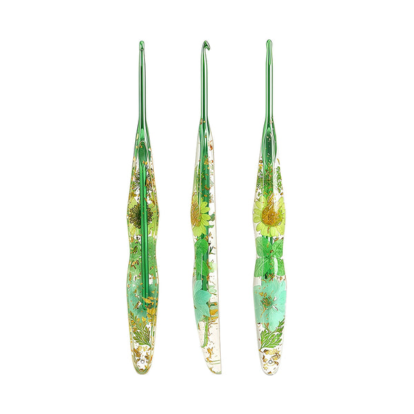 Three green-handled glass crochet hooks with dried flower and leaf inclusions, displayed vertically on a plain white background.