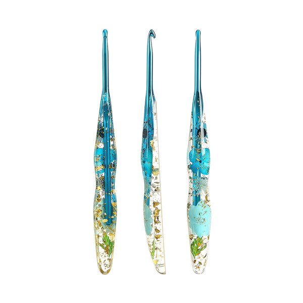 Three glass crochet hooks with blue handles and embedded gold foil and green botanical details, arranged vertically on a plain white background.