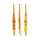 Three glass crochet hooks with amber-colored handles containing embedded dried flowers and green leaves, arranged vertically on a plain white background.