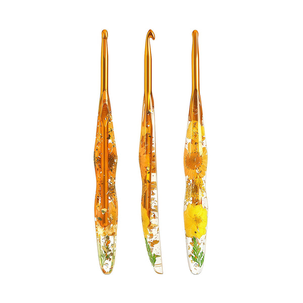 Three glass crochet hooks with amber-colored handles containing embedded dried flowers and green leaves, arranged vertically on a plain white background.
